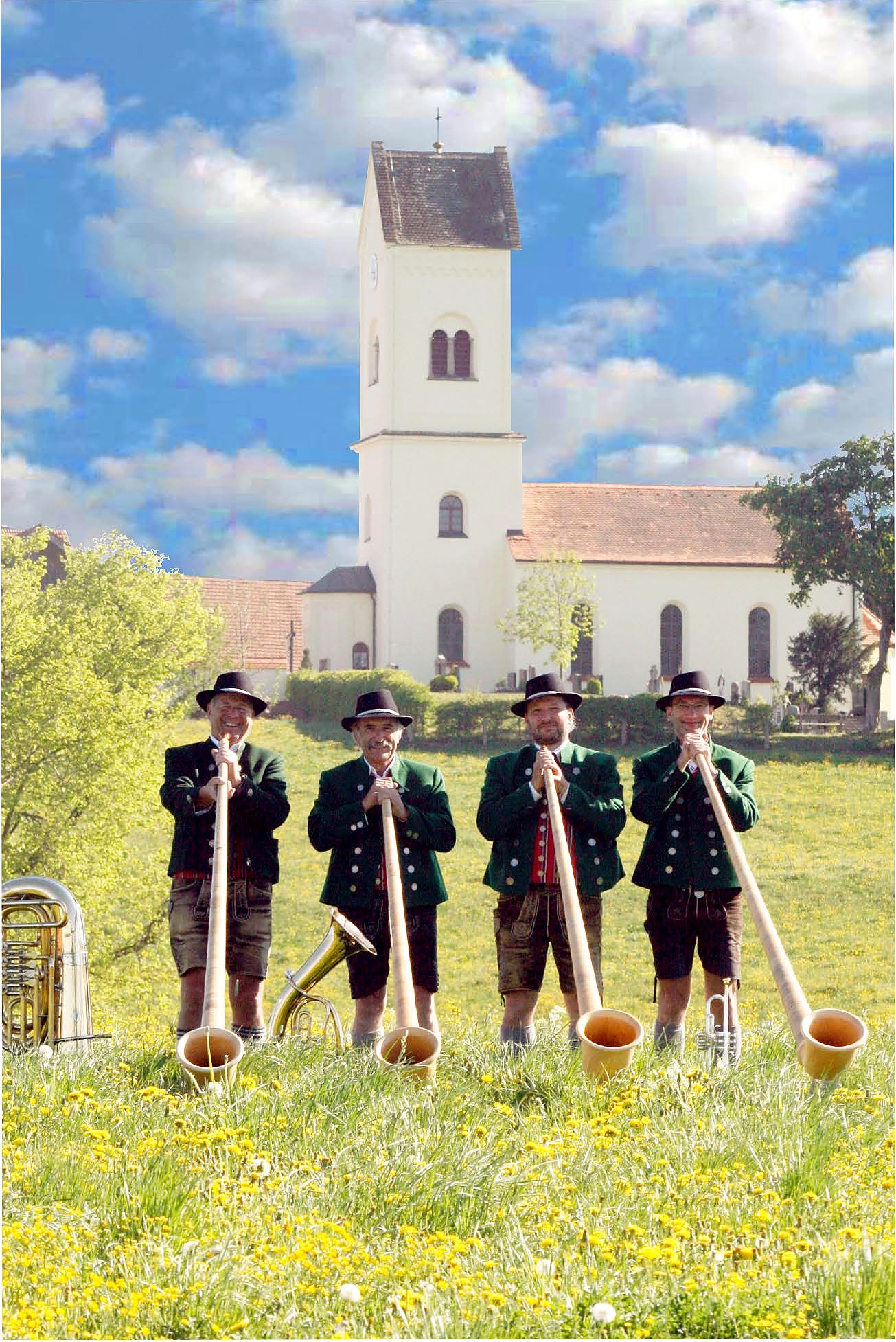 Vier Musiker in Tracht mit Alphörnern auf einer Blumenwiese vor der Kirche in Machtlfing